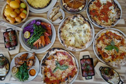 A spread of various pizzas, roasted vegetables, grilled chicken, and desserts on a wooden table.