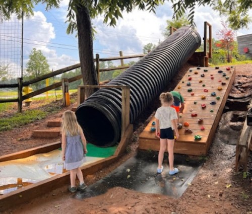 Children play on a playground with a large tube slide and a climbing wall, surrounded by trees and a sunny sky.