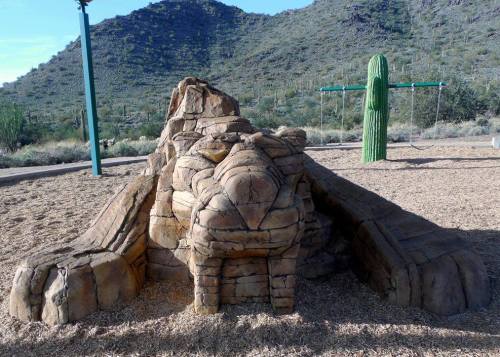 A rock formation resembling a lion sits in a playground, with a cactus and mountains in the background.