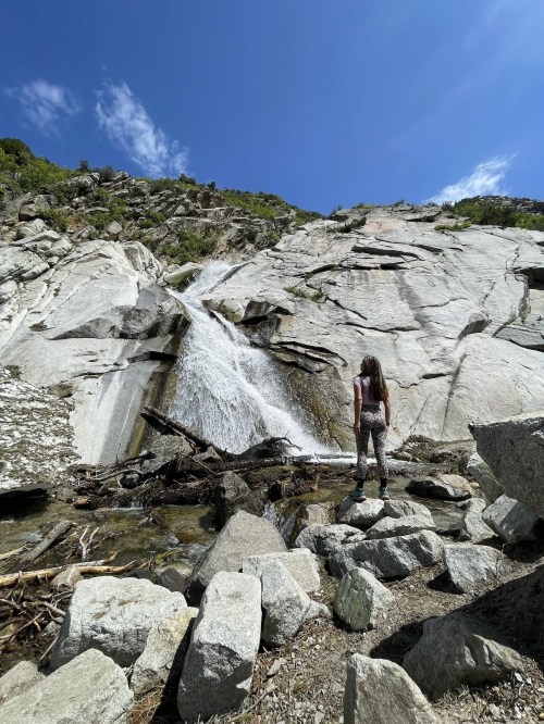 A person stands near a waterfall cascading down a rocky cliff under a clear blue sky.
