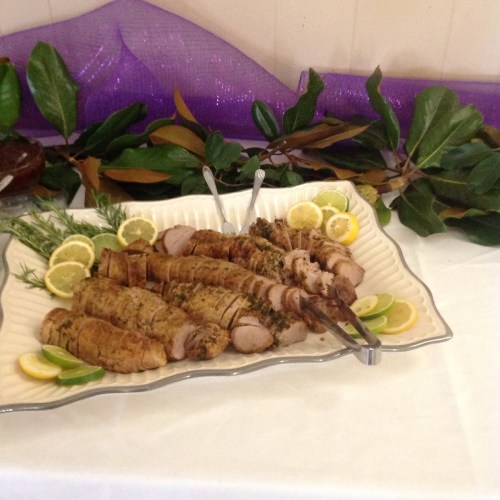 A platter of sliced roasted meat garnished with lemon slices and herbs, set on a table with decorative foliage.