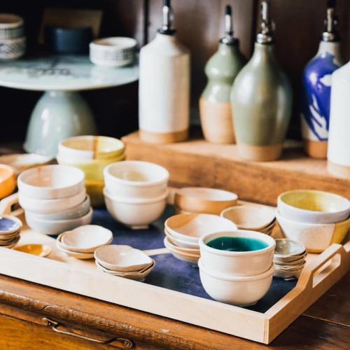 A wooden tray filled with various ceramic bowls and cups, surrounded by decorative bottles on a wooden shelf.
