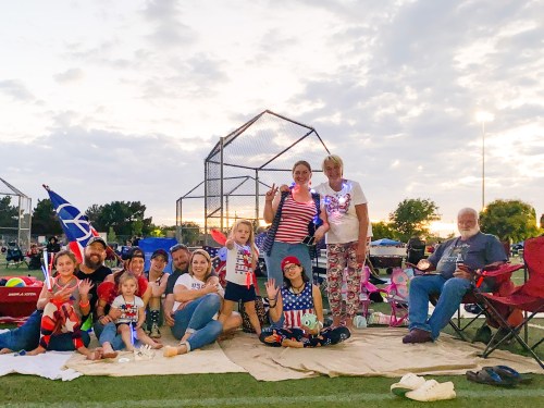 A group of people in patriotic attire sitting on a blanket at a park, smiling and waving, with a cloudy sky above.