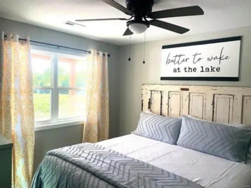 Cozy bedroom with a bed, decorative pillows, a ceiling fan, and a sign that reads "better to wake at the lake."