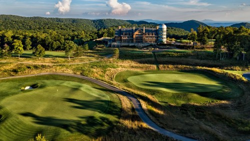 Aerial view of a golf course with rolling greens and a large lodge in the background, surrounded by trees and mountains.