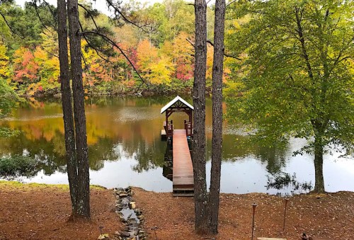 A serene lake view with a wooden dock surrounded by colorful autumn trees and tall pine trees.