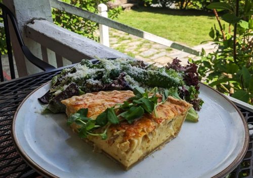 A slice of savory quiche on a plate, accompanied by a fresh salad topped with cheese, set outdoors on a sunny day.
