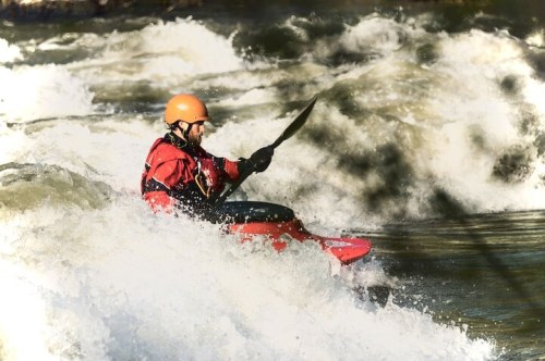 A kayaker in a red kayak navigates through turbulent whitewater rapids, wearing a helmet and paddling with determination.
