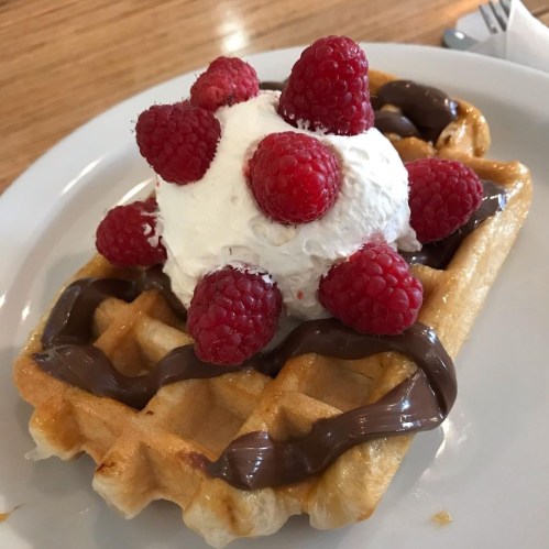 A waffle topped with whipped cream and fresh raspberries, drizzled with chocolate sauce on a white plate.