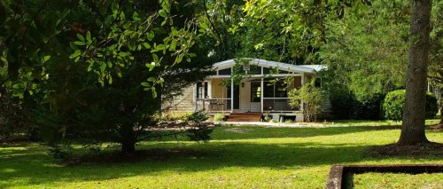 A cozy house surrounded by greenery, featuring a porch and large windows, set in a peaceful, sunny yard.