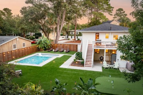 A spacious backyard with a pool, green lawn, and a two-story house surrounded by trees and seating areas.