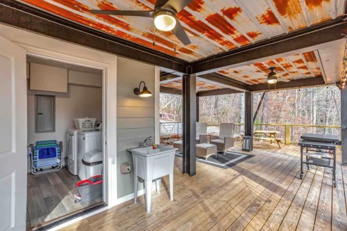 A covered porch with a rustic ceiling, featuring a laundry area and seating space overlooking a wooded view.