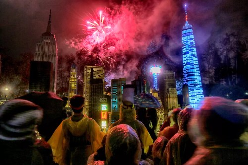 Crowd in raincoats watches fireworks over a city skyline at night, featuring iconic buildings and colorful lights.