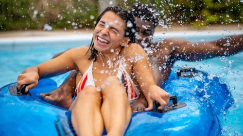 A joyful couple splashes water while sitting on a blue inflatable raft in a pool, enjoying a sunny day.