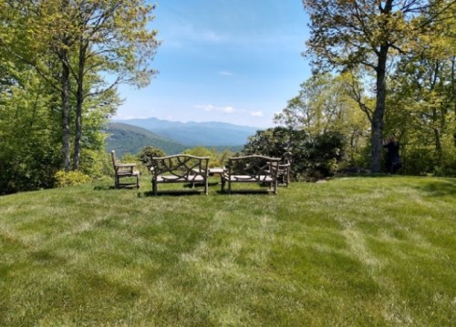 A serene outdoor scene with wooden chairs on a grassy area, overlooking distant mountains and a clear blue sky.