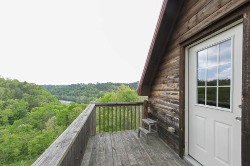 A wooden balcony with a door, overlooking a lush green landscape and river in the distance under a cloudy sky.