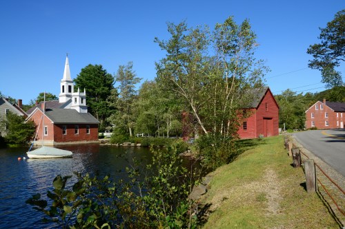 A serene scene featuring a church and red barn by a calm lake, with trees and a clear blue sky in the background.