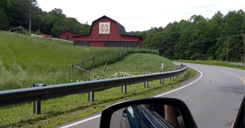 A red barn with a decorative star stands beside a winding road, surrounded by green fields and trees.