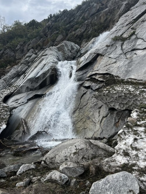 A waterfall cascading down rocky terrain, surrounded by boulders and sparse vegetation under a cloudy sky.