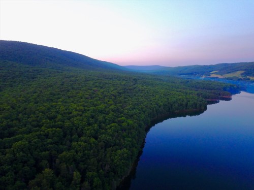 Aerial view of lush green hills surrounding a calm lake at dusk, with soft pastel colors in the sky.