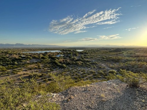 A scenic view of a desert landscape with shrubs, a lake, and mountains under a clear sky at sunset.