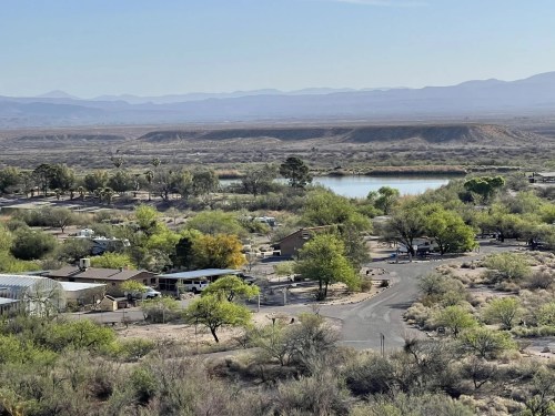 Aerial view of a desert landscape with a small lake, greenery, and scattered homes near winding roads.
