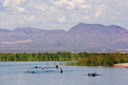 People swimming in a calm lake surrounded by lush greenery and mountains under a partly cloudy sky.