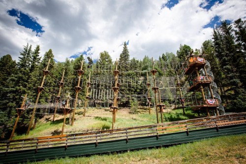 A high ropes course with wooden platforms and ropes, set in a forested area under a blue sky with clouds.