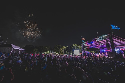 A lively outdoor concert scene with a large crowd, colorful stage lights, and fireworks in the night sky.