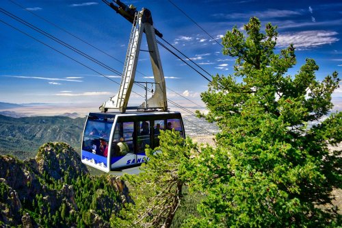 A cable car ascends a mountain, surrounded by lush greenery and a clear blue sky. Scenic views in the background.