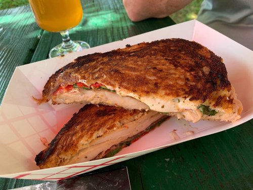 A toasted sandwich filled with chicken, peppers, and greens, served on a paper tray with a drink in the background.