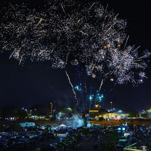 Colorful fireworks illuminate the night sky over a crowded parking lot, creating a festive atmosphere.