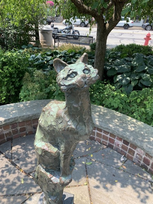 A bronze cat sculpture stands on a stone bench, surrounded by greenery and a glimpse of a street in the background.