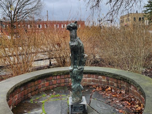A bronze statue of a bird perched on a stone pedestal, surrounded by bare shrubs and a cloudy sky.
