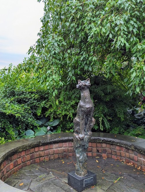 A bronze cat statue stands on a stone pedestal, surrounded by lush greenery and a circular brick wall.