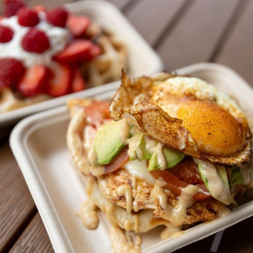 A close-up of a savory breakfast dish with a fried egg, avocado, tomato, and drizzled sauce on a plate. Waffles in the background.