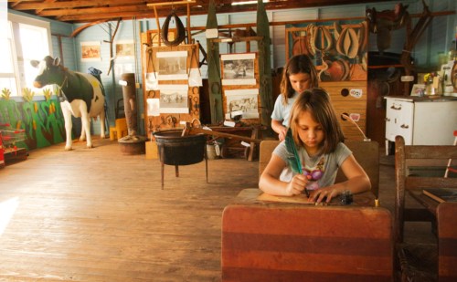 Two children engage in activities inside a rustic barn, with a cow statue and vintage farm tools in the background.