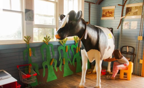 A child interacts with a life-sized cow model in a colorful room decorated with farm-themed murals.