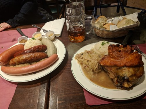 Two plates of hearty German food: sausages, sauerkraut, mashed potatoes, and a roasted pork knuckle, with beer and bread rolls.
