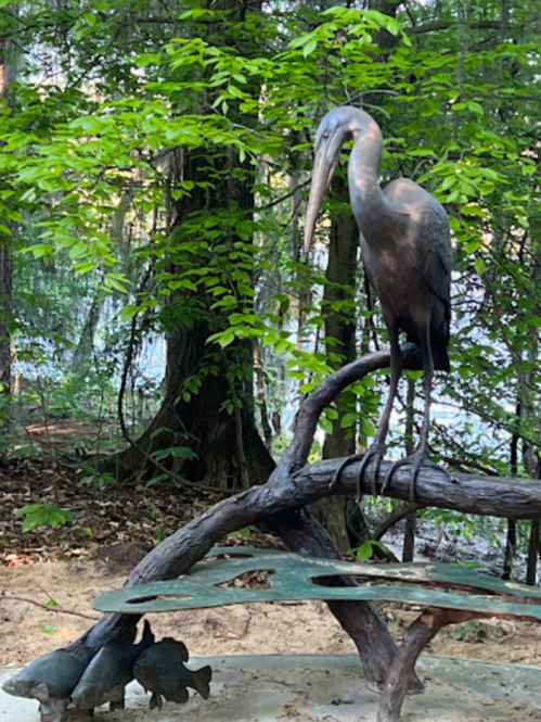 A heron sculpture stands on a branch, surrounded by greenery, with small animal figures beneath it.