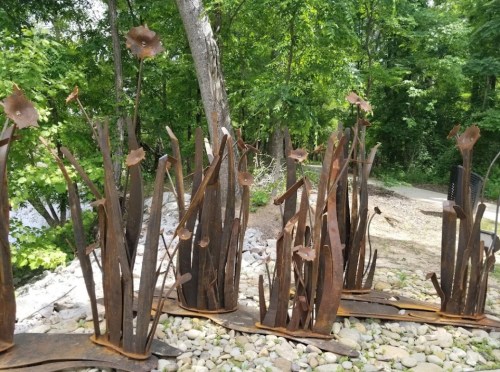 Rusty metal sculptures resembling tall plants and flowers, set among greenery and stones in a natural outdoor setting.