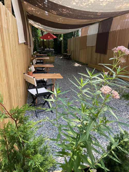 A serene outdoor café space with wooden tables, chairs, and greenery, shaded by fabric canopies.