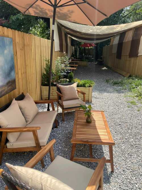 A cozy outdoor seating area with wooden furniture, plants, and an umbrella, surrounded by a gravel path and wooden fence.