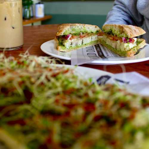 A close-up of a sandwich with green filling and vegetables, served on a plate with a drink in the background.