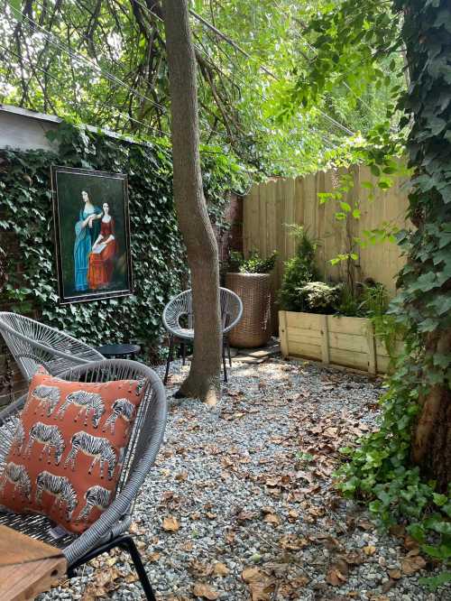 A serene garden space with a painting, two chairs, and greenery, surrounded by gravel and fallen leaves.