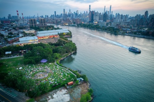 Aerial view of a riverside park with people relaxing, city skyline in the background, and a boat on the water.