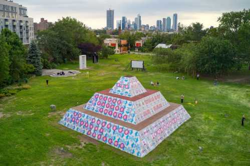 A colorful pyramid sculpture in a park, with a city skyline in the background and people enjoying the green space.