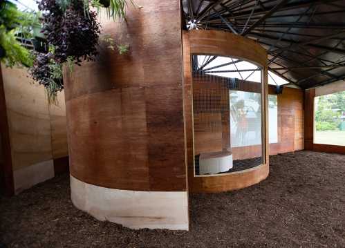 A modern wooden structure with a curved wall and large windows, surrounded by mulch and greenery.