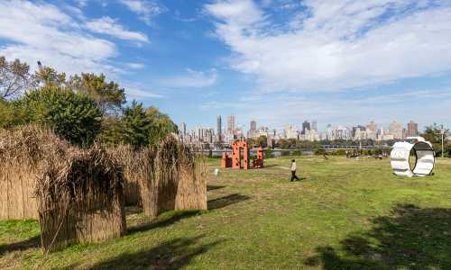 A park with sculptures and tall grass, overlooking a city skyline under a partly cloudy sky.