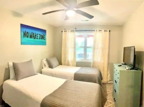 A cozy bedroom with two beds, a TV, and light curtains, featuring a "No Wake Zone" sign on the wall.
